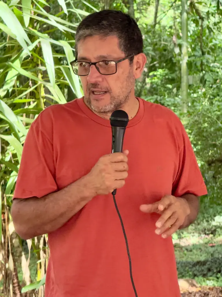 Hombre con gafas y camiseta naranja habla por un micrófono en un entorno natural.