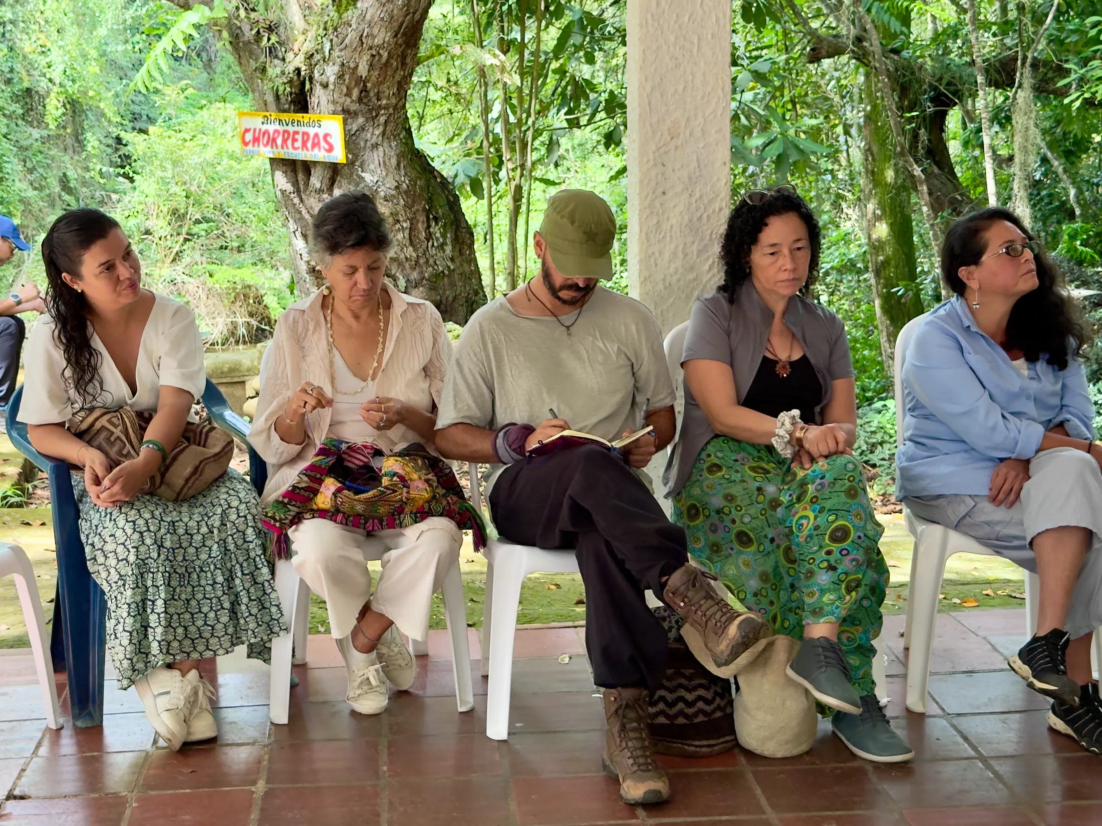 Grupo diverso de personas sentadas en sillas bajo un dosel, escuchando atentamente.